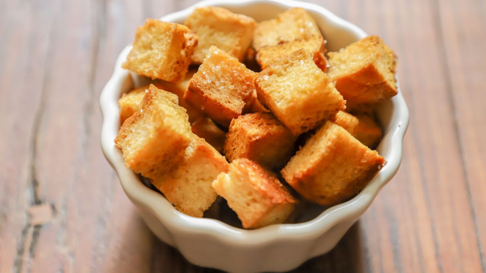 Slices of stale sourdough bread on a wooden cutting board next to a bowl of homemade croutons and a small jar of breadcrumbs